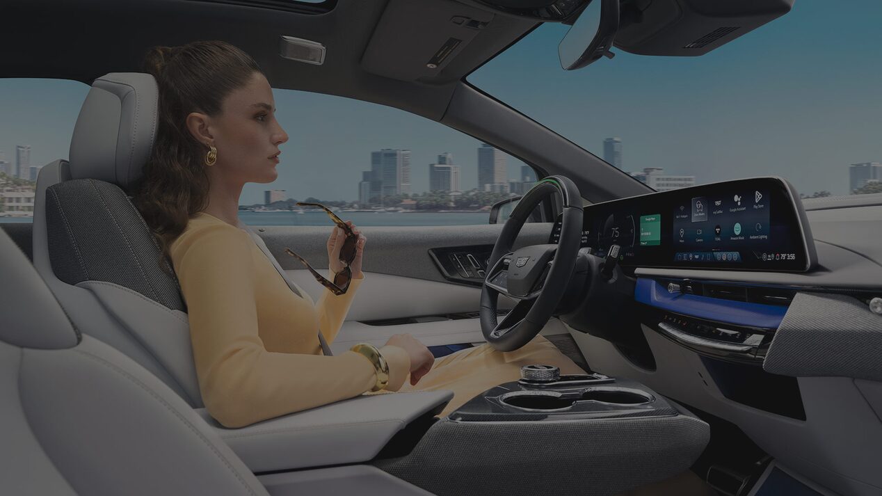 Passenger View of a Woman Driving a Cadillac OPTIQ Past the City While Utilizing Super Cruise Hands-free Technology