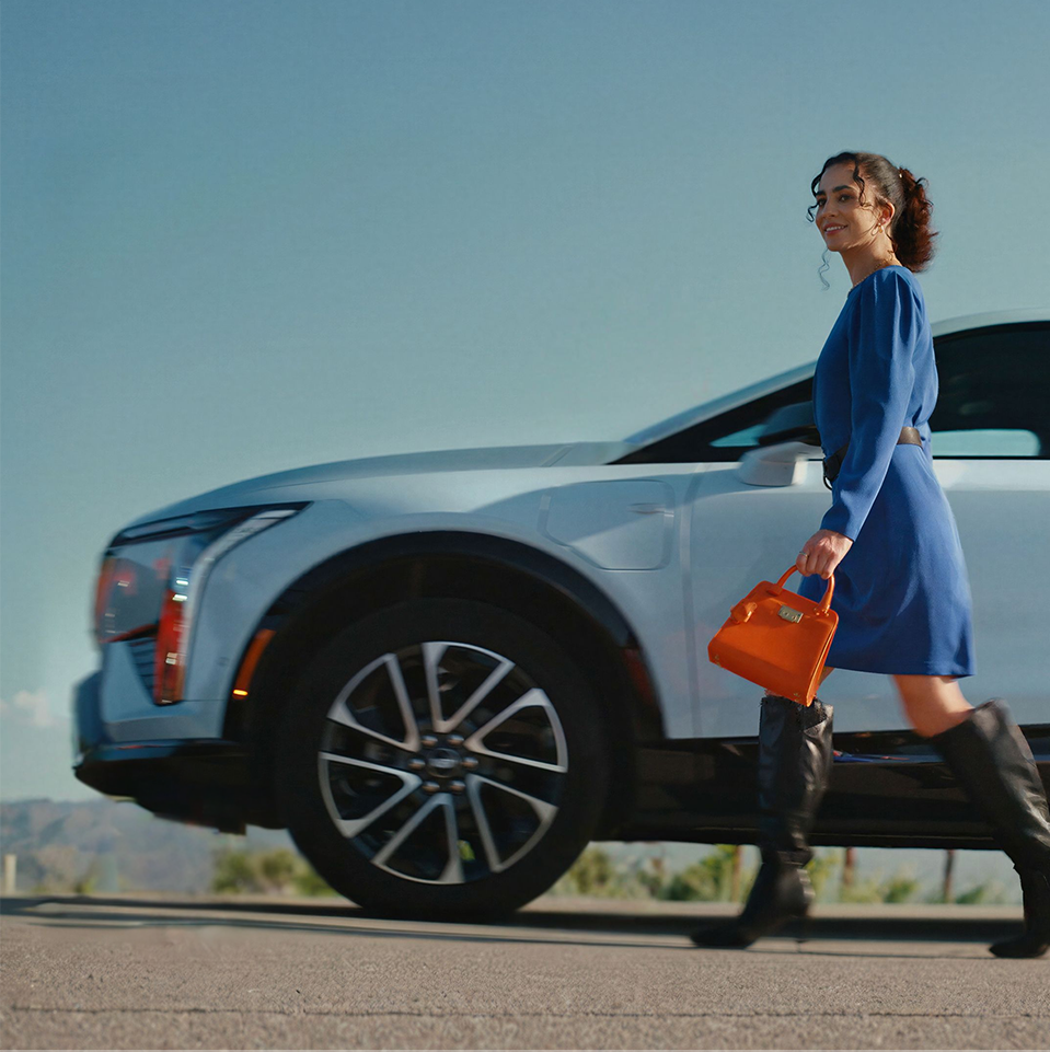 A Smiling Woman in a Blue Dress and Black Boots Walks Past a White Cadillac LYRIQ Holding an Orange Handbag
