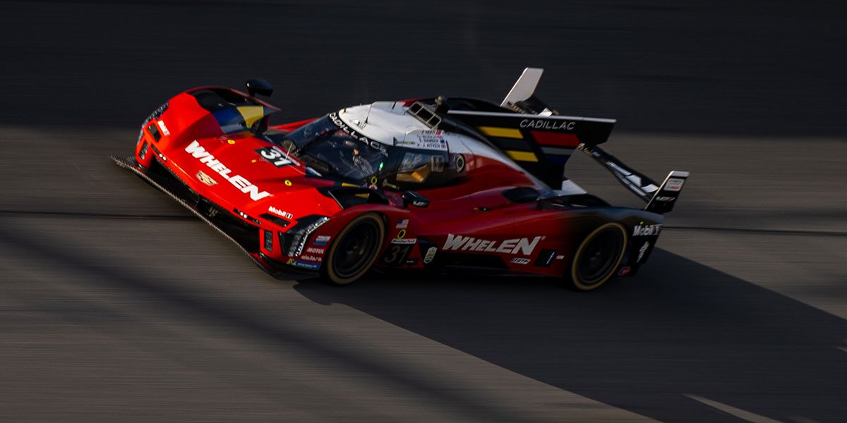 A Red Whelen Cadillac V-Series.R Race Car Races Down the Track at Dusk With Its Aerodynamic Body Low and Fast Against the Asphalt.