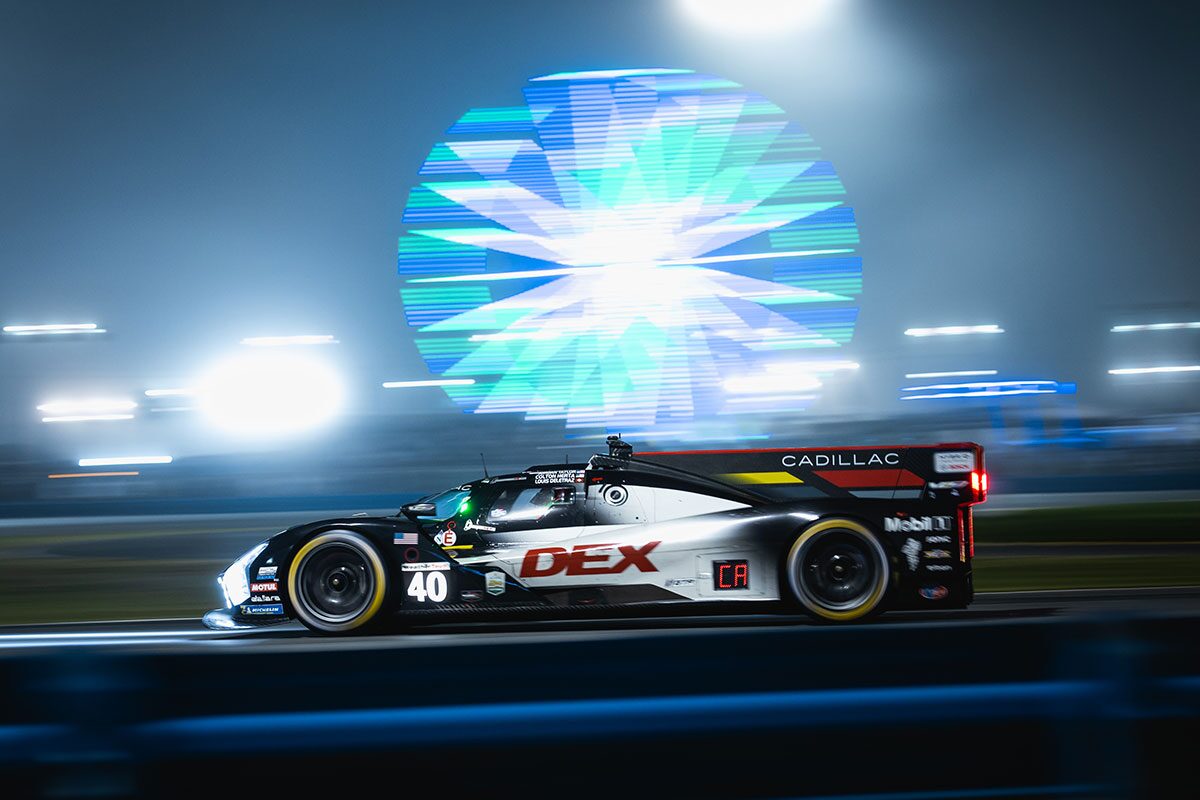 A Whelen Cadillac V-Series.R Race Car Speeds Along the Track at Night With a Large Multicolored Ferris Wheel Glowing Brightly Behind It.