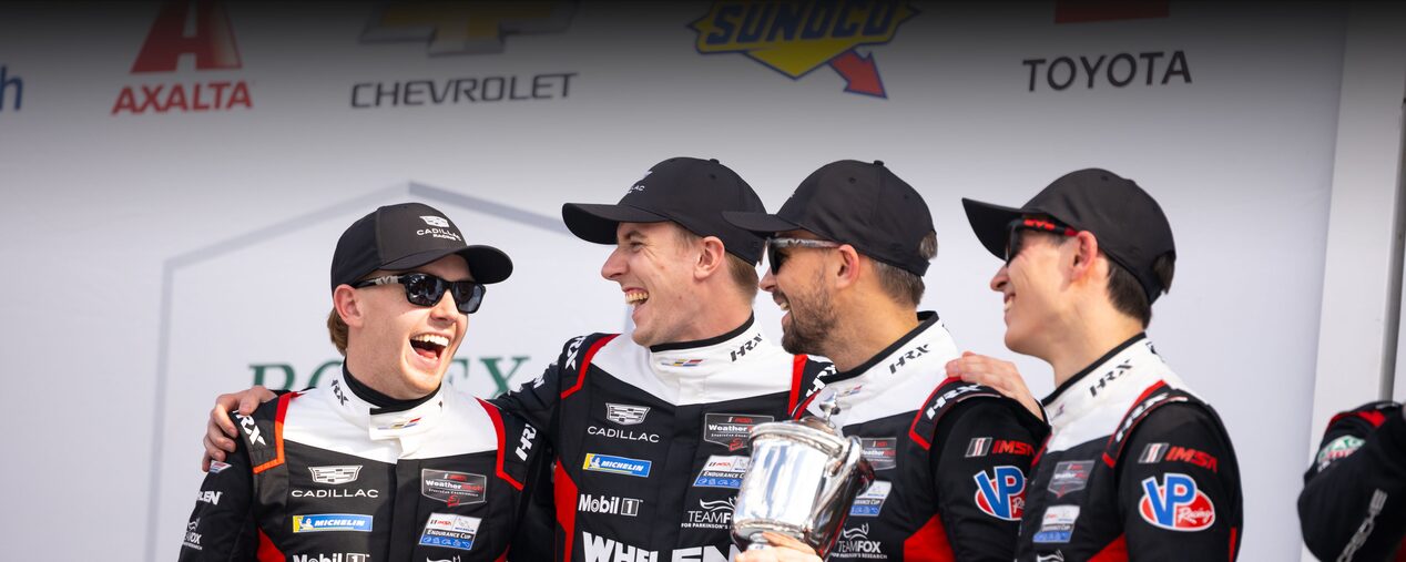 Four Happy Cadillac Race Car Drivers in Black and White Suits and Sunglasses Celebrating on a Podium With a Silver Trophy.