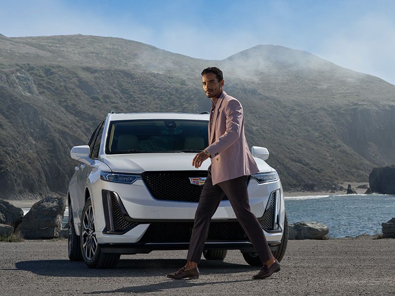 A Man Looking at the Camera While Walking in Front of the Cadillac XT6 with a Stunning View of Mountains in the Distance