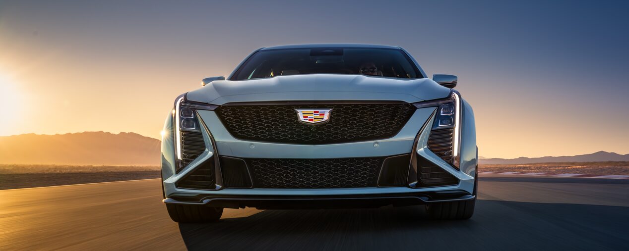 Low Front View of a Light Blue 2025 Cadillac CT5-V Driving Down the Road Next to a Setting Sun