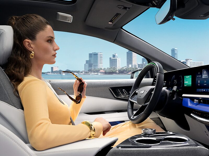 Passenger View of a Woman Driving a Cadillac OPTIQ Past the City While Utilizing Super Cruise Hands-free Technology